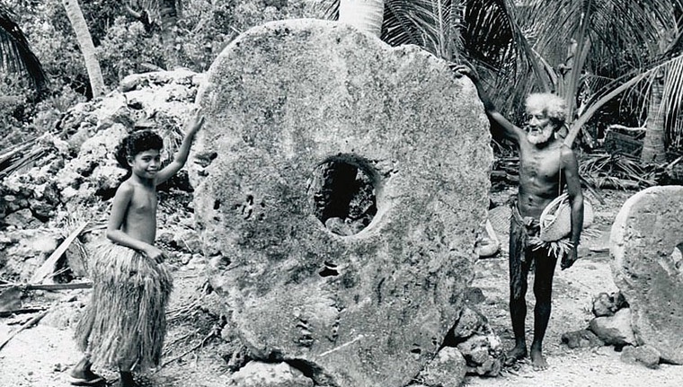 Historic photo of two Yap Islanders standing next to a large Rai stone, an ancient stone currency used in Micronesia, symbolizing wealth and traditional payroll practices.